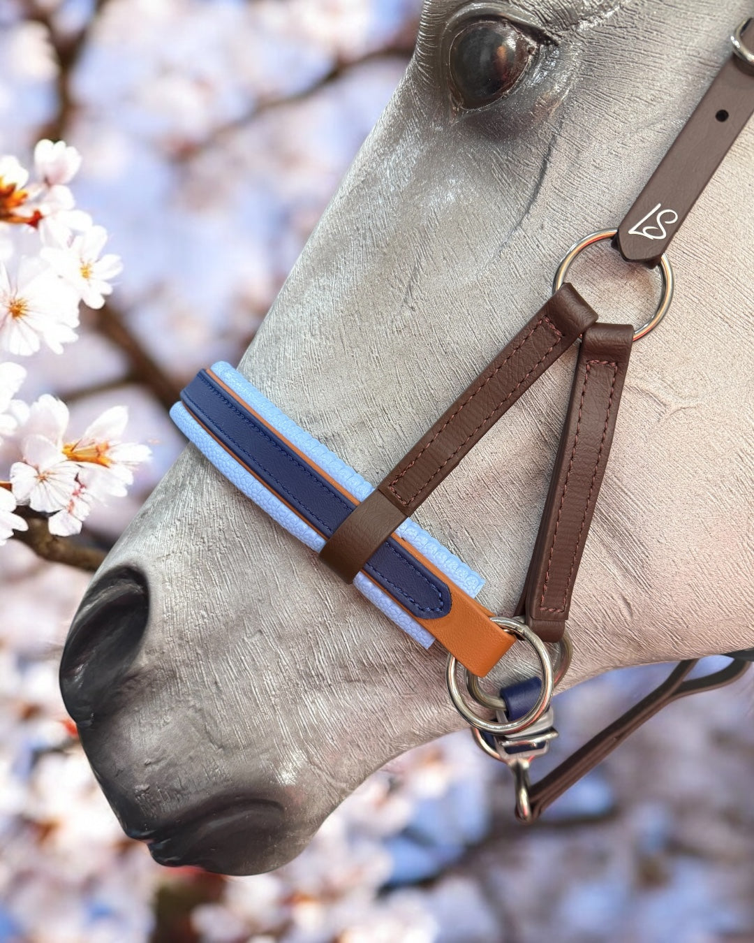 A gray horse wearing an LS Equestrian SidePull Bitless Bridle Option 2 with custom brown and blue accents is framed by cherry blossoms. The visible eye and muzzle contrast softly with the natural blossoms.