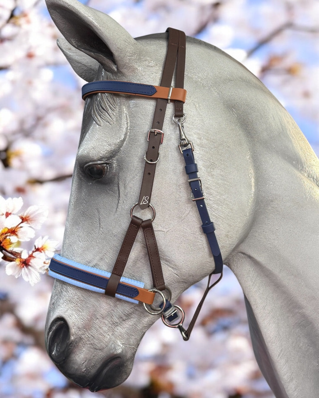 A gray horse stands gracefully against blooming white flowers, wearing a custom-made LS Equestrian SidePull Bitless Bridle Option 2 in BioThane®️ with blue and brown accents.