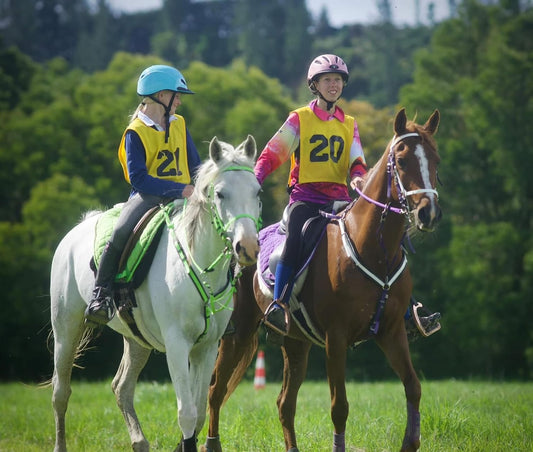 Two equestrians wearing numbered vests and helmets, part of the LS Equestrian NZ Starter Enduro Package, ride a white and a brown horse side-by-side through a grassy field with trees and a striped marker in the distance.