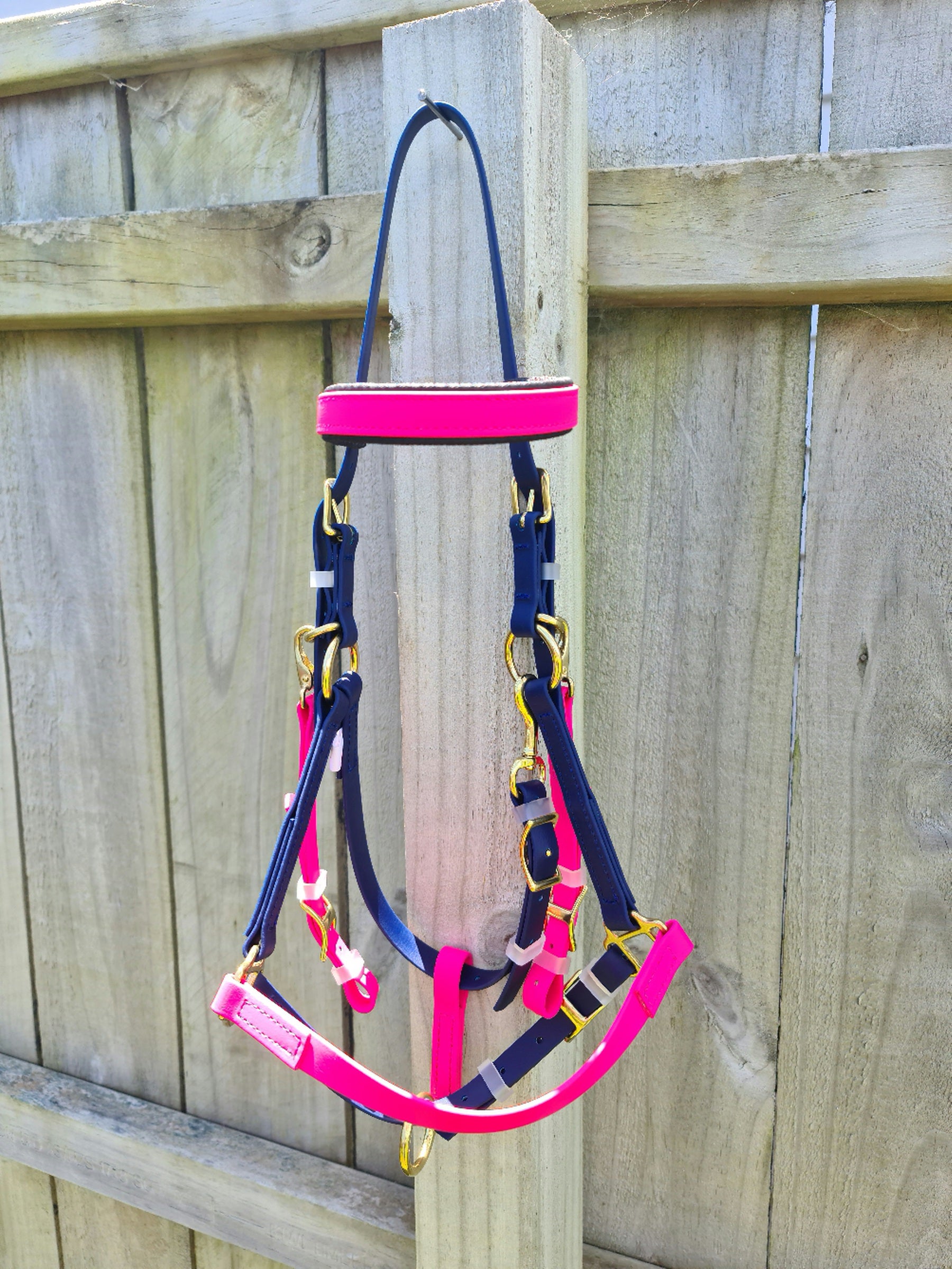 A LS Equestrian Bridle - Halter Bridle Navy & Pink, featuring gold-colored hardware, hangs on a wooden fence post. The weathered fence, with its visible grain patterns, contrasts beautifully with the new and neatly arranged halter. Nearby, endurance bridles hint at the rider's preparation for long trails.