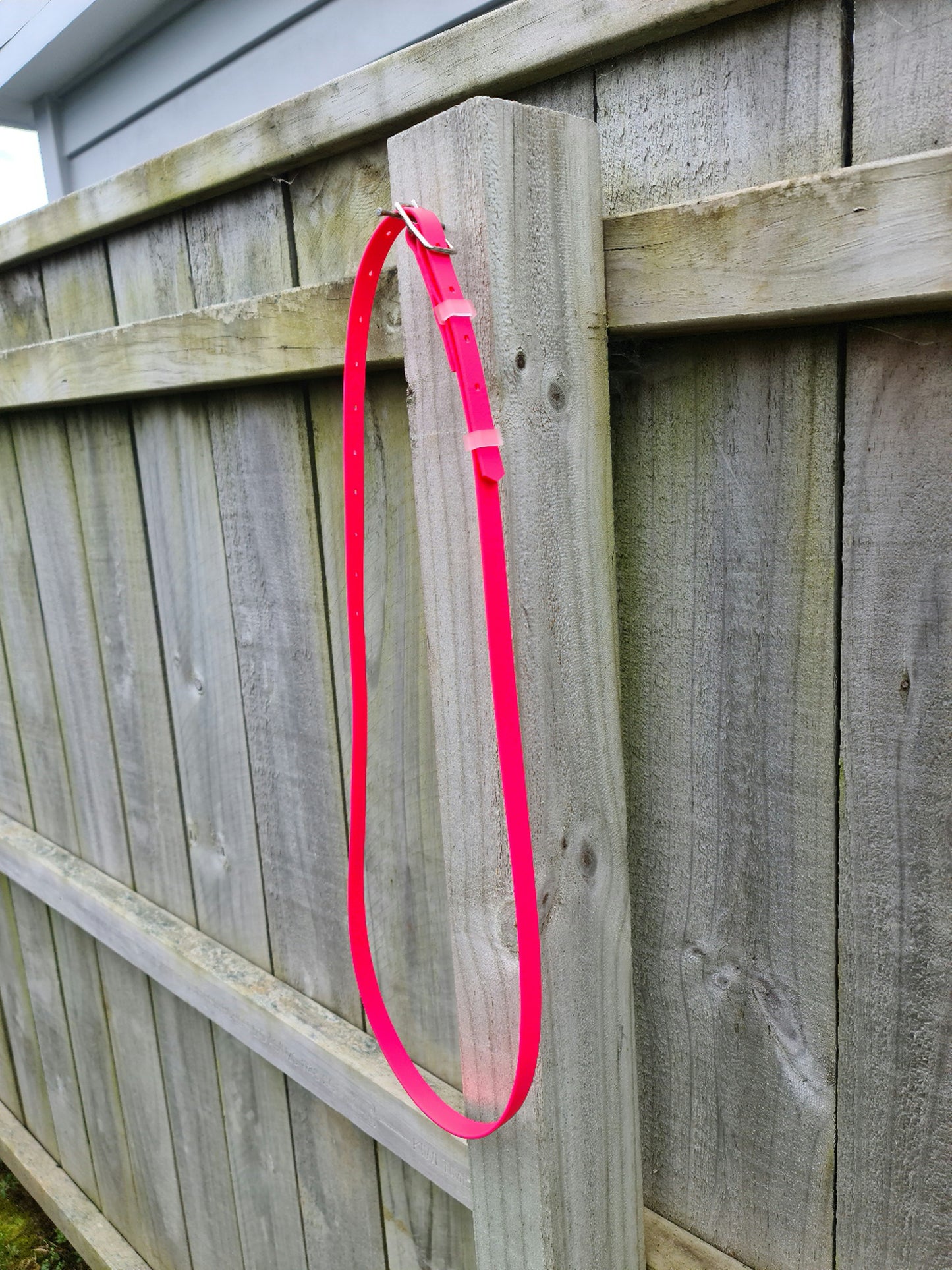 An "Oh Shoot" Neck strap from LS Equestrian made of vibrant pink, transparent BioThane material is hanging on a gray wooden fence. The buckle is fastened, and the neck strap appears slightly curved against the vertical post of the fence. The background shows the top edge of the fence with some visible sky.