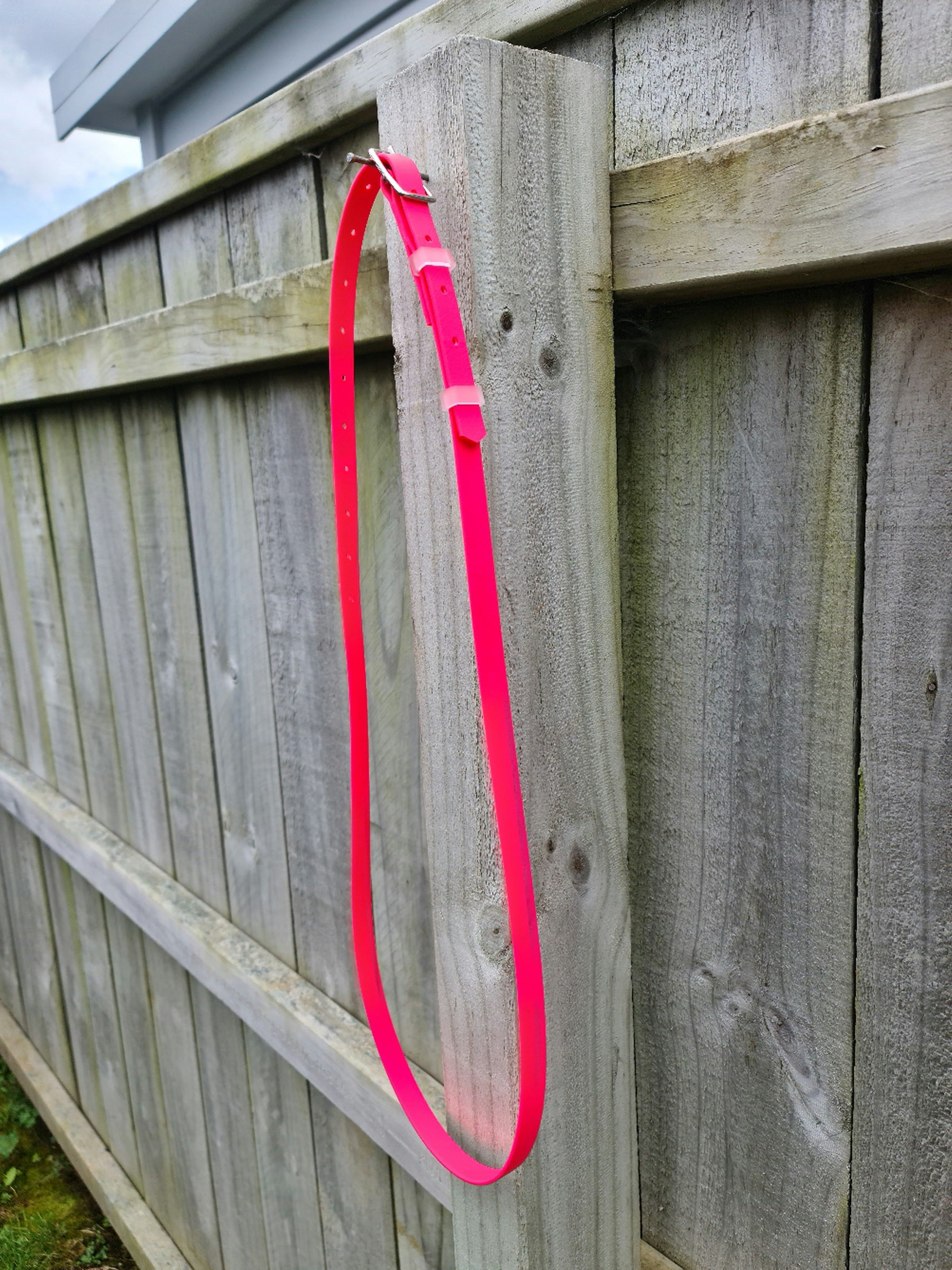 A bright pink "Oh Shoot" Neck strap by LS Equestrian hangs from the top of a wooden fence, resembling the vibrant tack often seen on bridles. The neck strap is long and loops back around itself, with the buckle visible at the top. The background features a portion of the fence and a grassy area below.