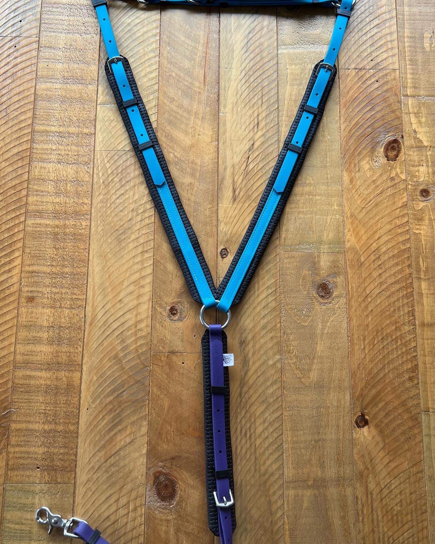 A V-shaped Biothane ®️ Breastplate - Design your own from LS Equestrian lies on a wooden surface, embodying the durability found in endurance bridles. The breastplate features blue and purple padded straps with black linings and silver hardware, including rings and buckles. A silver clasp is seen towards the bottom left corner of the image.