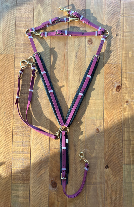 A maroon Martingale from LS Equestrian is laid out on a wooden surface. The harness, featuring black padding and brass fittings, showcases an elegant look enhanced by its BETA finish, with its straps, buckles, and rings neatly arranged. Note: Breastplate not included in the price.